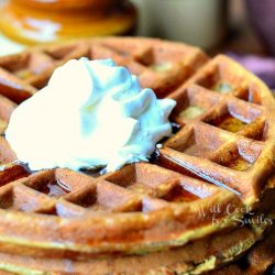 stack of gingerbread waffles with a whipped topping on small plate with fork at the bottom right of plate on a brown placemat. 2 brown and tan jars in background to the left