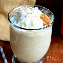 1 glass dessert mug with handle holding gingersnap milkshake with whipped topping on a wood table with strawas scattered around glass and brown cloth in background to the left