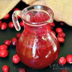 glass syrup jar filled with cranberry syrup on a black table with cranberries scattered around jar and brown tablenapkin in background at the top of the picture