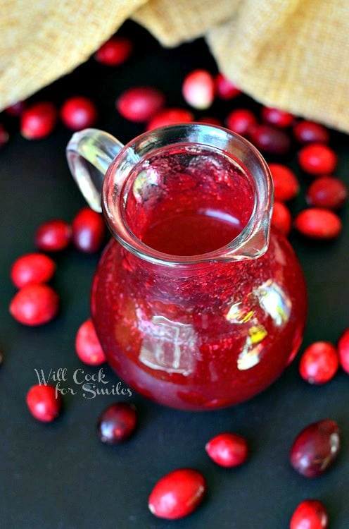 Cranberry Syrup in a glass serving pitcher with cranberries around it 