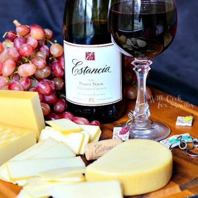 glass of red wine on cutting board with grapes and several different types of cheese and bottle of wine next to cheese