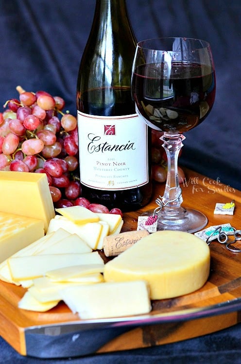 a glass of red wine with the bottle behind it on a a cutting board with cheese and grapes 