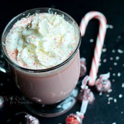 above view of clear glass coffee mug filled with peppermint white chocolate hot cocoa topped with whipped topping as peppermint hershey kisses and a candy cane are scattered at bottom of coffee mug