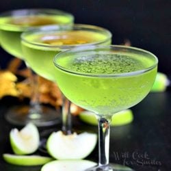 3 coctail martini glasses filled with sour apple champagne coctail with apple slices scatteredaround the bottom of the glasses and fall decor in the background