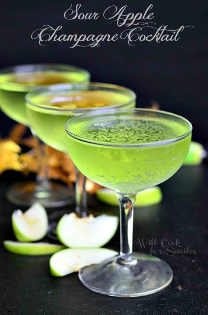 3 coctail martini glasses filled with sour apple champagne coctail with apple slices scatteredaround the bottom of the glasses and fall decor in the background