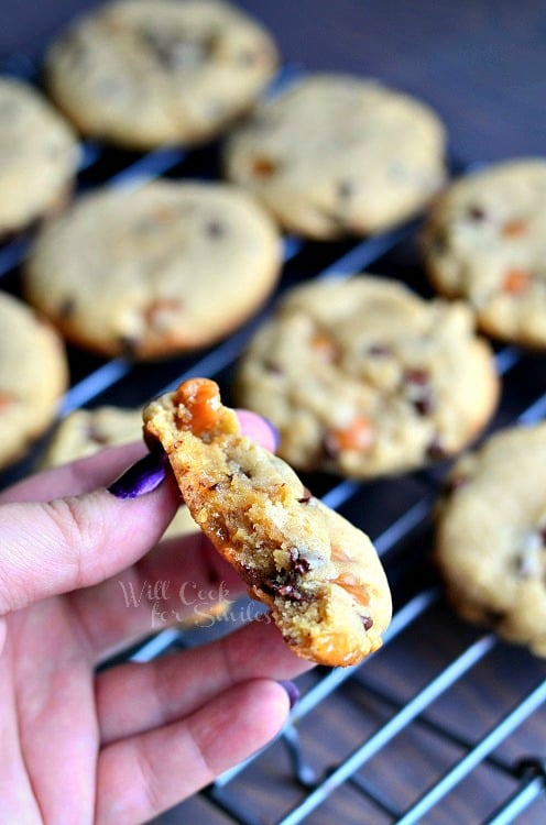 holding a Sumbitch Peanut Butter Chocolate Caramel Cookie and the rest of the cookies on a cooling rack in the background 