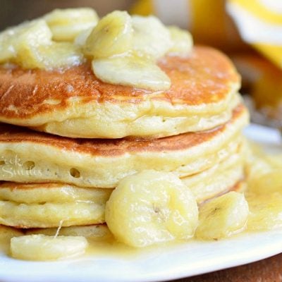 stack of bananas foster pancakes on white plate on wood table with yellow and white cloth in background
