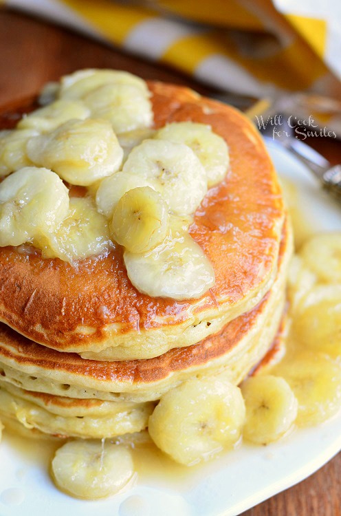 view from above of stack of bananas foster pancakes on white plate on wood table with yellow and white cloth in background