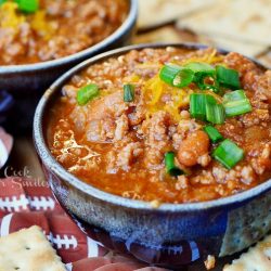 close up view of 2 small bowls of boozy chili on a blue platter with cracker scattered around bowls. Chili topped with shredded cheese and chives