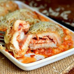 rectangular plate with chicken parmesan roll ups and fork holding one bite above first rollup. Plate is on a brown placemat on a wood table.