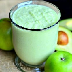 clear glass coffee mug filled with green apple avocado green smoothie on a burlap placemat with sliced avocado and apples to the right and background of glass