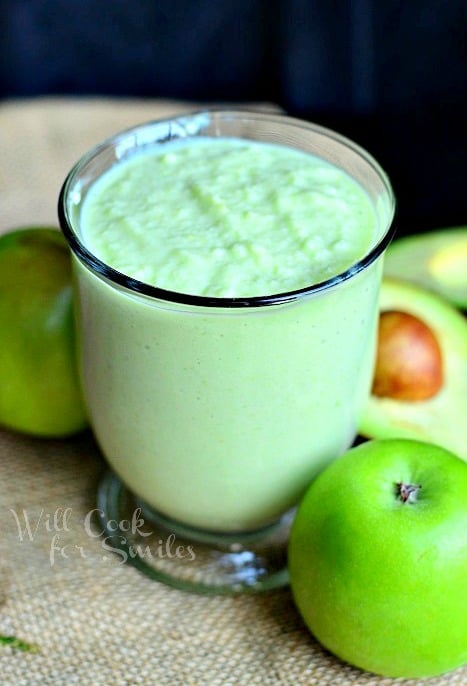 clear glass coffee mug filled with green apple avocado green smoothie on a burlap placemat with sliced avocado and apples to the right and background of glass