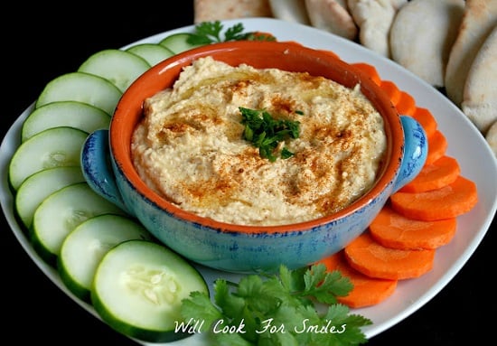 Hummus in a blue bowl on a white plate with cucumbers and carrots around it 