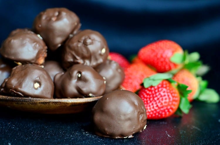 (No Bake) Chocolate Covered Peanut Butter Cheesecake Bites in a wood bowl and one on the table top with strawberries to the right 