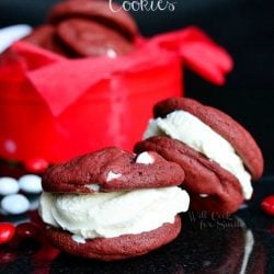 close up view of red velvet ice cream sandwich cookies on a black table with red and white candies scattered around cookies and red and white gift box in background