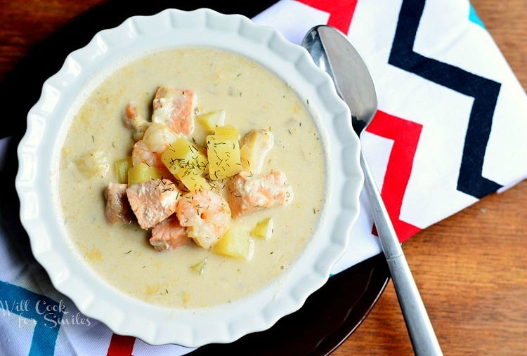 view from above white decorative bowl filled with seafood chowder on a white cloth with ziig zag colors on a black plate all on wood table with a spoon to the right of the bowl