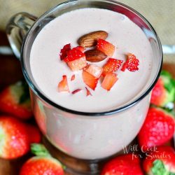 clear glass coffee mug filled with chocolate covered strawberry protein shake on a wood table with strawberries and straws around glass at bottom. Shake is topped with diced strawberries