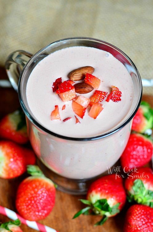 clear glass coffee mug filled with chocolate covered strawberry protein shake on a wood table with strawberries and straws around glass at bottom. Shake is topped with diced strawberries