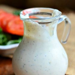 syrup jar with handle filled with homemade buttermilk ranch on a wood table with a garden salad in a white bowl in background to the left and a brown cloth to the right