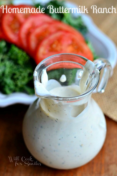 Homemade Buttermilk Ranch is served in a glass pouring dish. It's white in color. Behind the dressing is a dish of greens and slices of tomato.