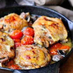 Roasted chicken thighs with tomatos and mushrooms on a black skillet on a wood table with a brown napkin in background