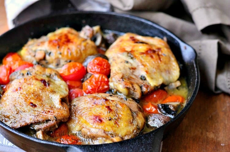 horizontal image of four baked chicken thighs in a cast iron skillet with tomatoes and mushrooms