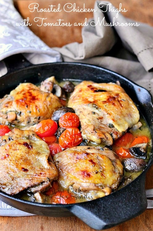 four baked chicken thighs in a cast iron skillet with tomatoes and mushrooms and brown napkin next to it