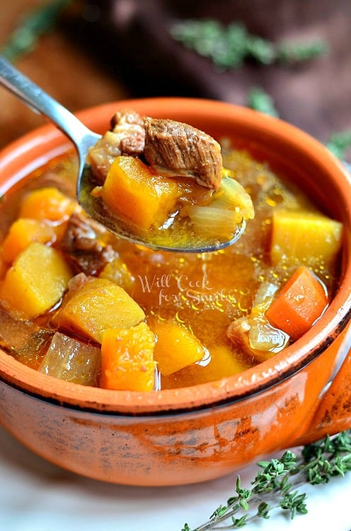 above view of winter squash beef stew in a red clay bowl on a white plate with a spoon holding portion of soup above the bowl