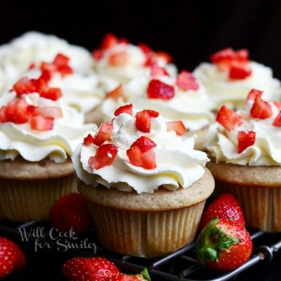 several strawberry cupcakes with mascarpone frosting on cooling rack above black table with strawberries scattered below