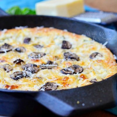 close up view of asiago mushroom onion pizza baked in skillet on a blue cloth with cheese and cheese grater at top of picture