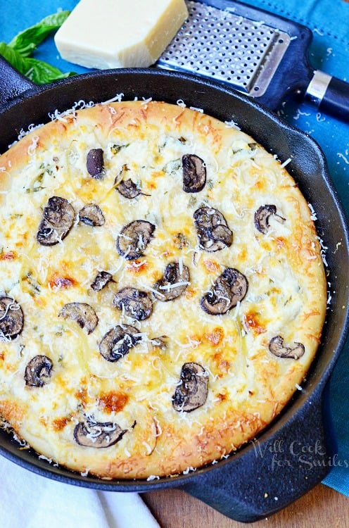 Asiago Mushroom & Onion White Pizza Skillet in a cast iron skillet with a blue table clothe and cheese and cheese grater above it 