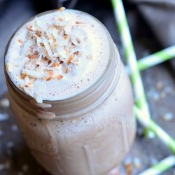 close up view of mason jar filled with coconut mocha milkshake on a grey table with green and white straws scattered around jar and a greyish cloth in background