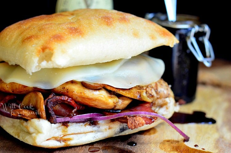 Chicken sandwich with mushrooms, bacon, and red onions, with a guinness glaze on a table with a guinness beer and a jar of glaze in the background