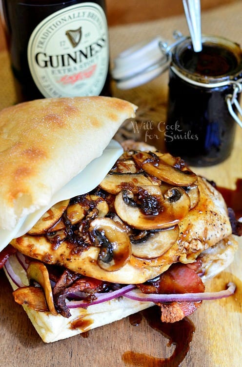 Chicken sandwich with mushrooms, bacon, and red onions, with a guinness glaze on a table with a guinness beer and a jar of glaze in the background