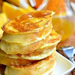 stack of orange creamsicle french toast slices on a white plate on wooden table with yellow and white cloth in background with syrup jar