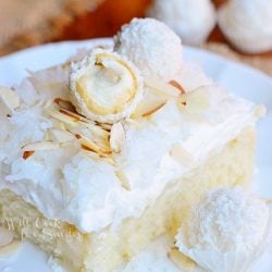 1 slice of rafaello poke cake coconut cake with white chocolate coconut cream and whipped topping on a small white plate on a brown burlap placemat on a wood table with Raffaello candy in background
