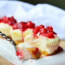 rectangular white plate with 3 skinny mini strawberry cheesecakes with strawberry topping lined up with fork on left side of plate on a red table cloth with white cloth in background