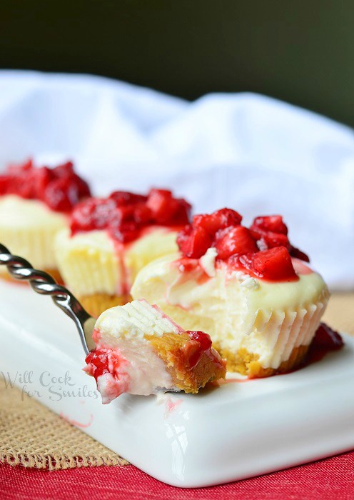 rectangular white plate with 3 skinny mini strawberry cheesecakes with strawberry topping lined up with fork on left side of plate on a red table cloth with white cloth in background