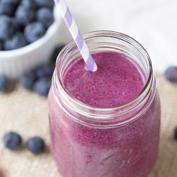 above view of mason jar glass filled with berry and toasted coconut smoothie on a brown placemat with berries in a small white crock in background to the left and additional berries scattered at base of mason jar