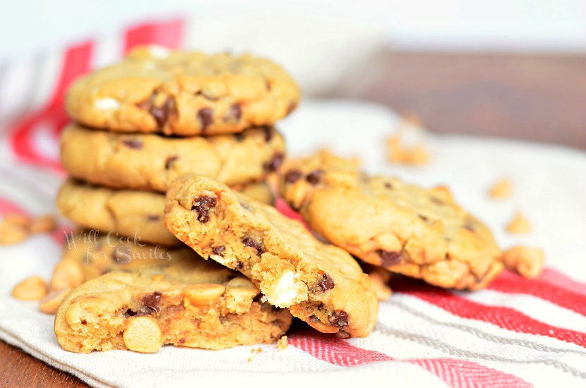 Peanut Butter Cookies that are loaded with white chocolate chips, mini chocolate chips and peanut butter morsels stacked up on a white and red kitchen towel with one cut in half