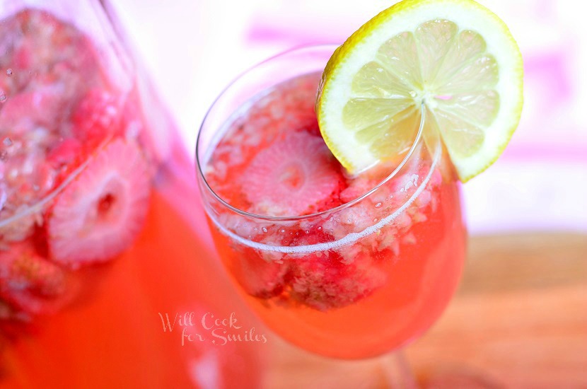 beverage jug filled with pink berry sangria on a wood plank with a wine glass filled with sangria to the right of jug