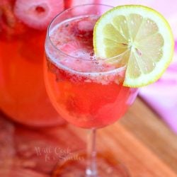 beverage jug filled with pink berry sangria on a wood plank with a wine glass filled with sangria in foreground