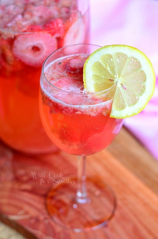 beverage jug filled with pink berry sangria on a wood plank with a wine glass filled with sangria in foreground