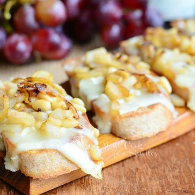 several Presidents cheese crostinis lined up on a wood plank on wood table with grapes, bottle of wine and cheese package in background