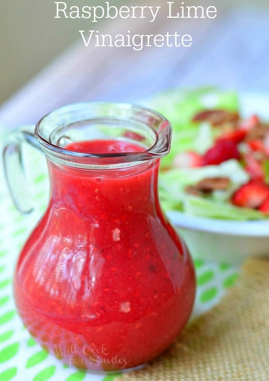 Raspberry Lime Vinaigrette is served in a glass pouring dish. It's red in color and the seeds are visible. Behind the vinaigrette, is a bowl of salad with fresh fruit and nuts. The bowl of salad fades into the background.
