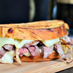 close up of steak and onion grilled cheese sandwich on a wood plank in front of a clear glass beer mug filled with stout beer and a bottle of beer in background