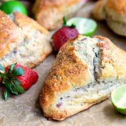 close up view of 1 strawberry key lime scones with strawberries and cut limes in foreground on a piece of brown wax paper and several more scones and fruit in background