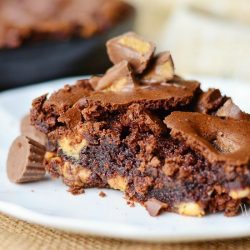 1 portion of ultimate peanut butter cup brownie on a small white plate on brown placemat with whole brownie dish in background to the left