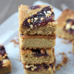 4 salted peanut butter and jelly blondies stacked on a white paper towel on wood table with more blondies below