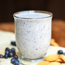 glass mug filled with blueberry muffin milkshake on a burlap cloth on wood table with blueberries and cookies scattered along the bottom of the glass